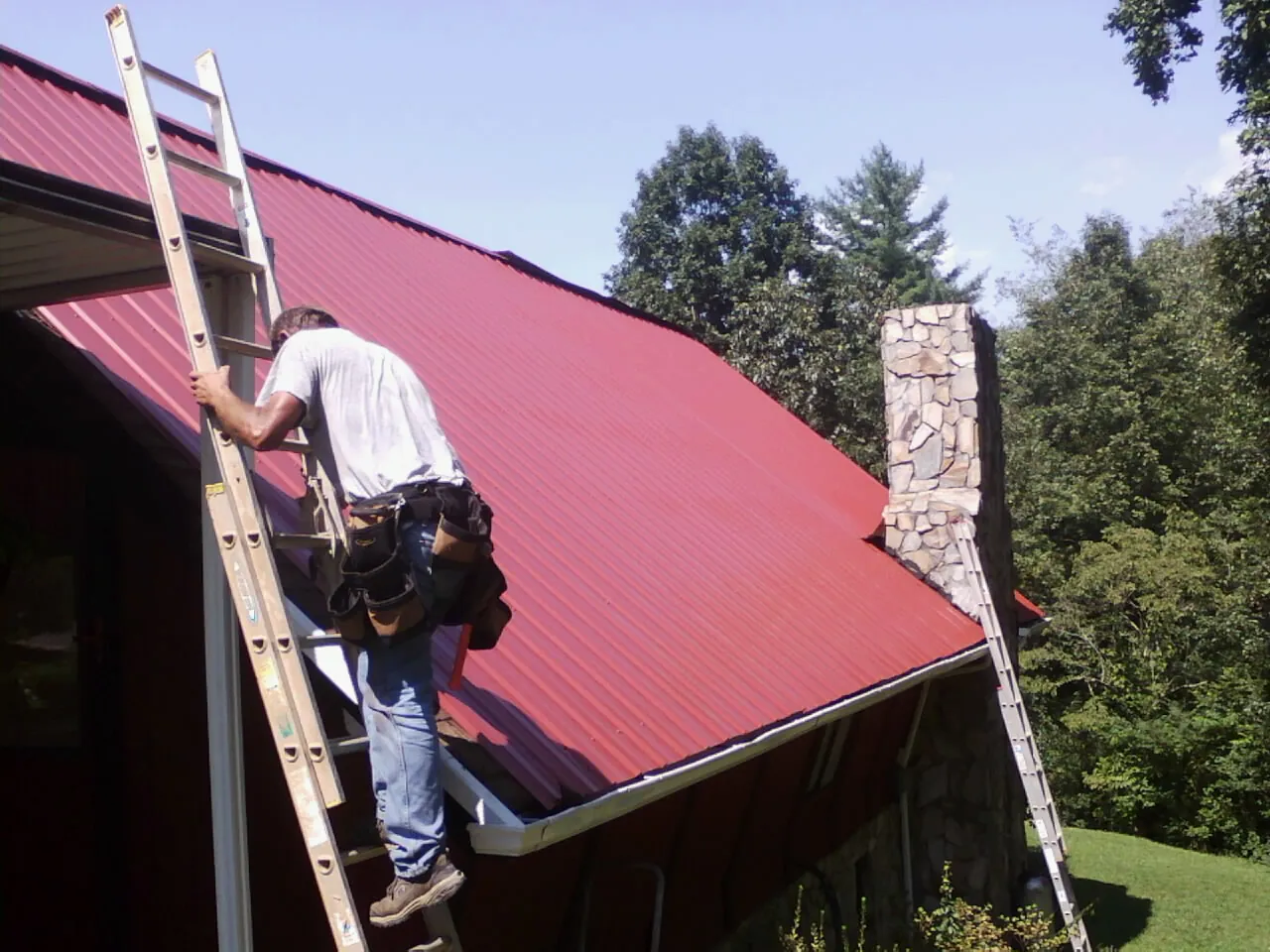 Roofer on a ladder beside a red metal roof and stone chimney — Soffit & Fascia Repair by Roofing Brothers in Kennebunk